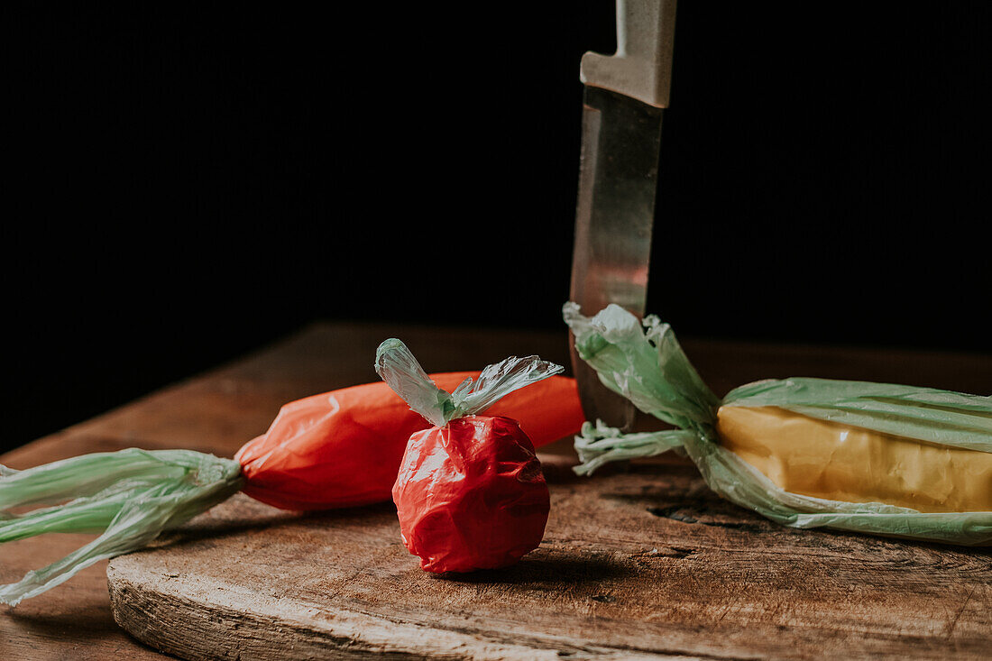 Artistic display of plastic bags shaped like a red tomato, an orange carrot, and a yellow bell pepper beside a chef's knife on a chopping board