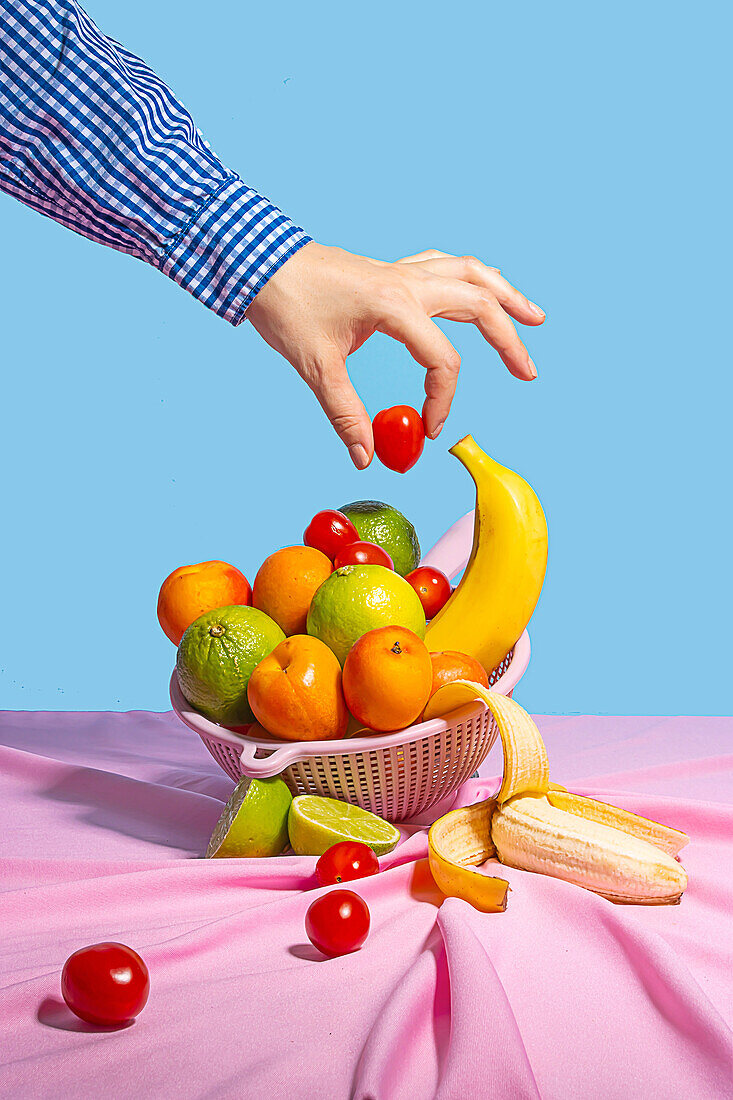 Anonyme Person, die eine Kirschtomate aus einer Plastikrutsche mit frischem Obst auf einem Tisch mit rosa Tischtuch nimmt