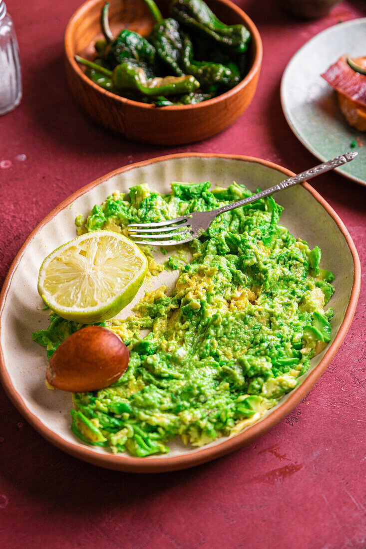 Delicious vegetarian smashed avocado guacamole garnished with lime and onion on plate with fork during cooking process