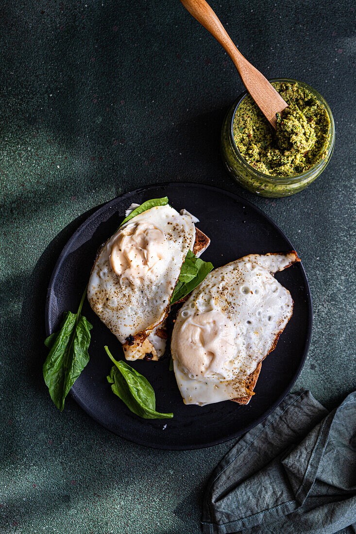 Top view of black plate with delicious homemade wholegrain toasts with fresh green spinach, fried egg and pesto sauce placed on gray table