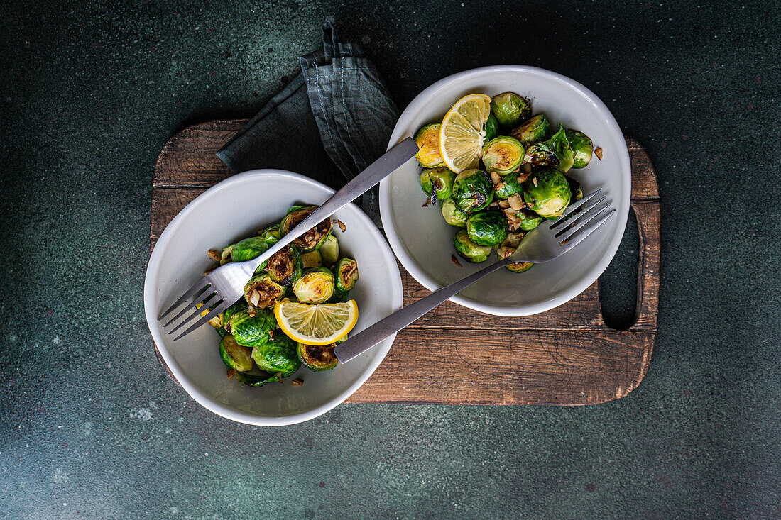 Top view of bowl of grilled Brussels sprouts seasoned with garlic and spices, garnished with a slice of lemon, served on a wooden board with a fork