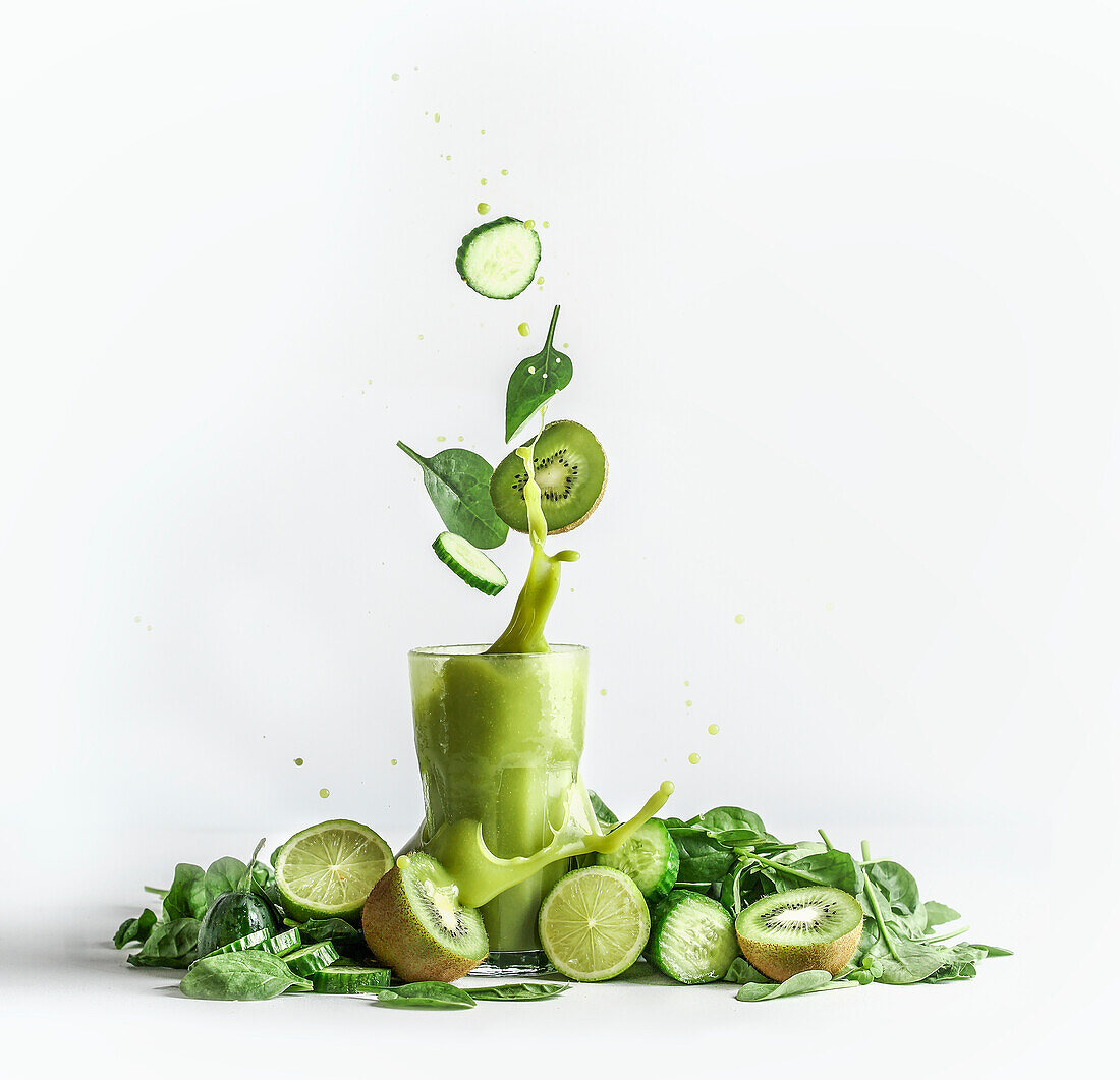 Splashing of green smoothie in glass with and flying ingredients: cucumber,kiwi and spinach leaves at white background with heap of green fruit and vegetable. Healthy refreshing drink