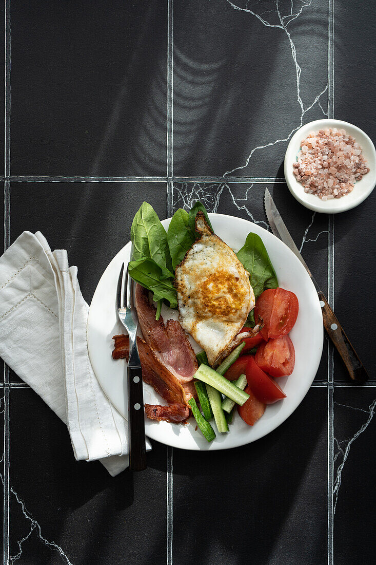Top view of well balanced breakfast setup with a fried egg, bacon strips, and assorted vegetables like tomato, cucumber, and spinach on a white plate over a dark marble background