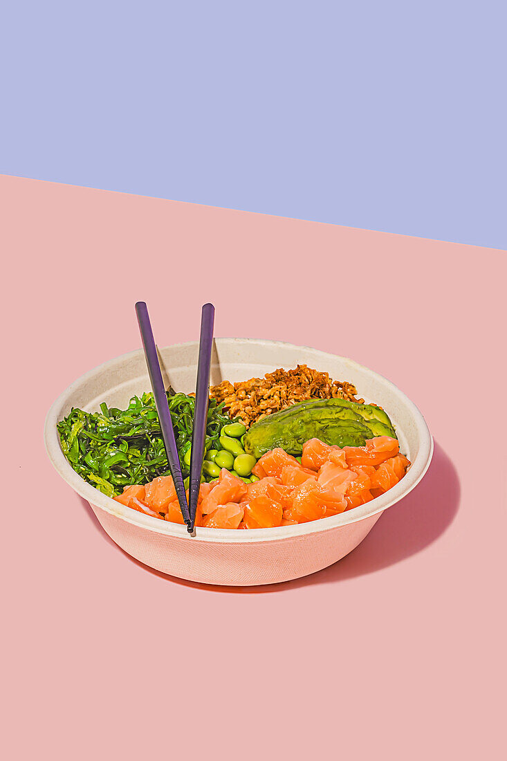Schale mit frischem Lachs, grünen Edamame-Bohnen, knackigem Algensalat, cremiger Avocado und knusprigem Müsli, begleitet von einer leichten Dip-Sauce und Stäbchen, vor einem zweifarbigen pastellfarbenen Hintergrund