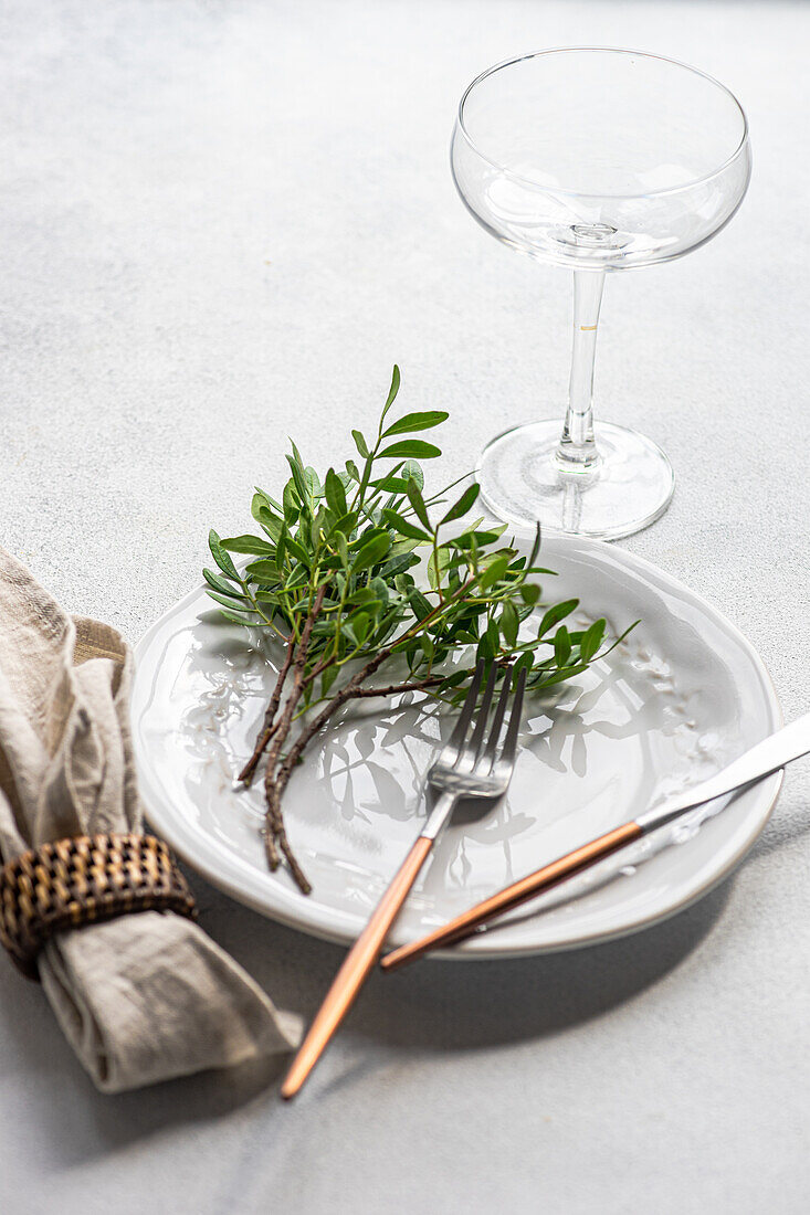 Hoher Winkel einer Tischdekoration mit einer frischen Pistazienpflanze auf einem Teller mit Besteck neben einer Serviette und einem Glas auf einer grauen Fläche im Tageslicht