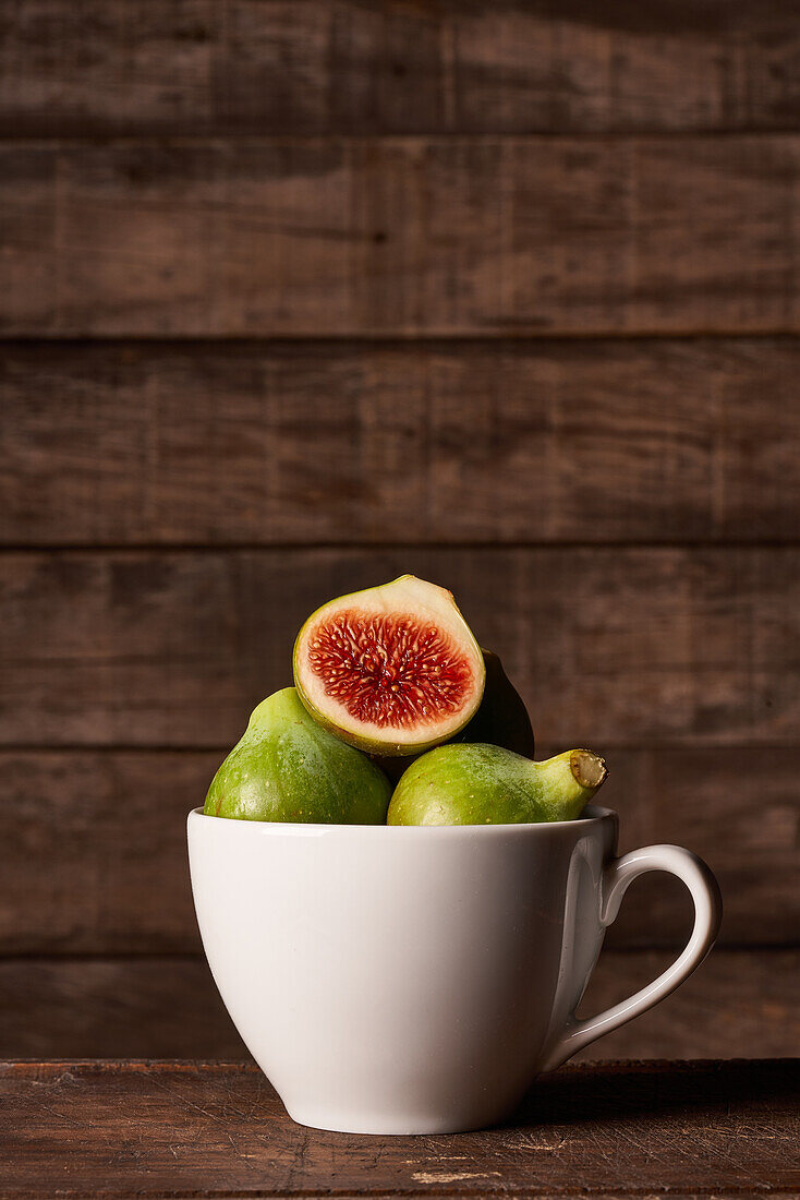 A rustic still life of fresh green figs, one sliced open to showcase the red interior, paired with a wooden honey dipper, all beautifully arranged in a white ceramic cup set against a dark wooden backdrop.