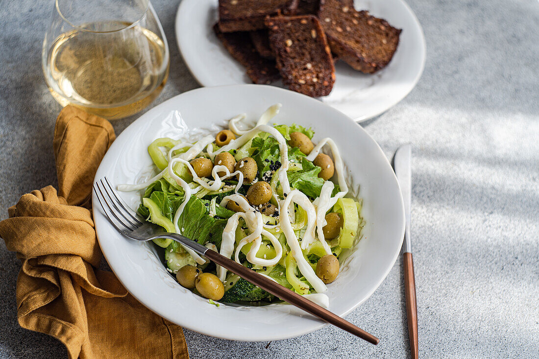 High angle of vegetable salad with green vegetables like lettuce, cucumber, olives, green bell pepper with homemade cheese and sesame seeds placed near plate of bread, glass of wine and cutlery against gray background