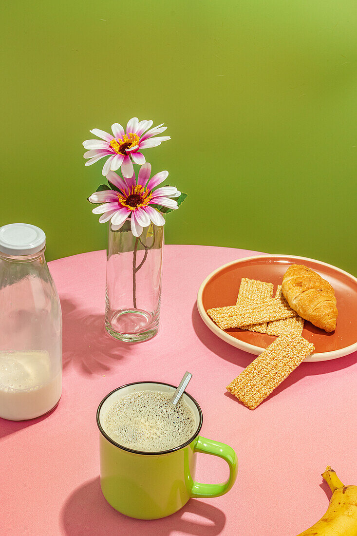 Appetitanregende Croissants und Banane neben Glasflasche mit Milch und Wasser mit Zitronenscheibe vor Blumen in Vase und Tasse mit aromatischem Kaffee auf rosa Tisch vor grünem Hintergrund