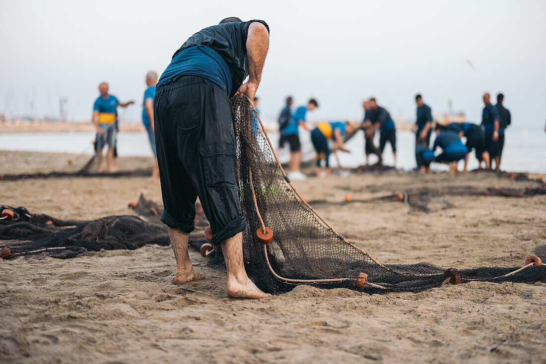 Rückenansicht eines anonymen Mannes in nasser Kleidung, der nach unten blickt, während er am Sandstrand ein Fischernetz trocknet, vor dem sich unscharfe Fischer mit ihrem Fang versammelt haben