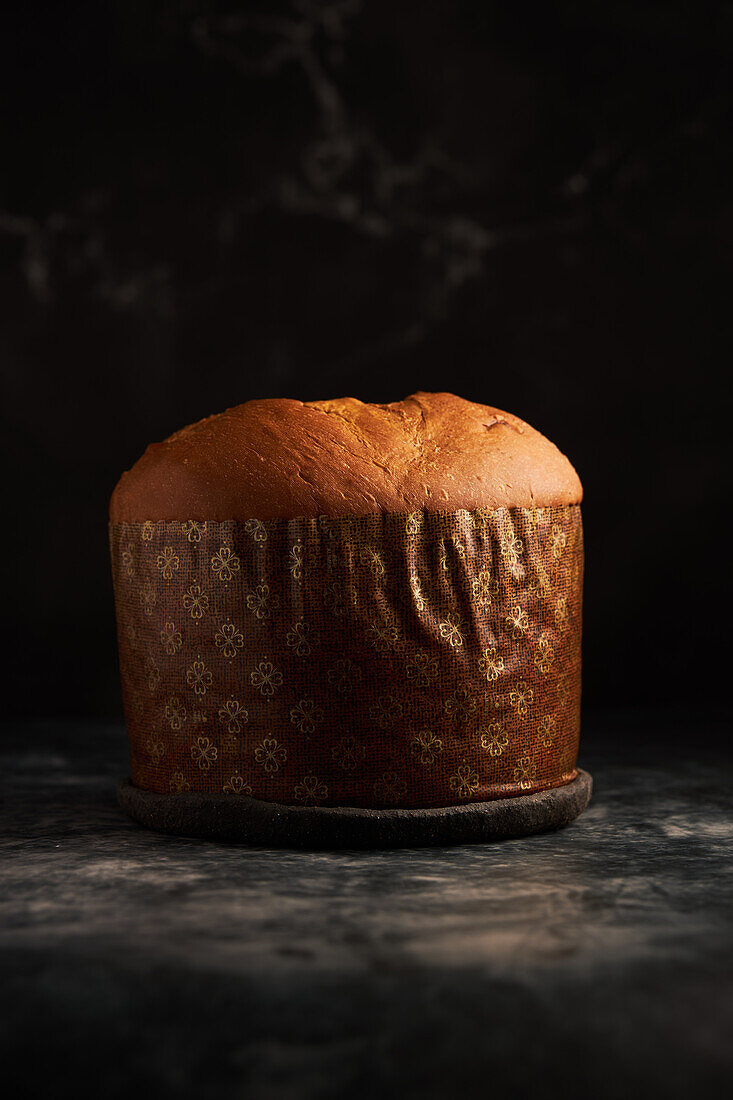 A freshly baked panettone, with a rich golden crust, sits against a moody dark backdrop, emphasizing its warmth and texture