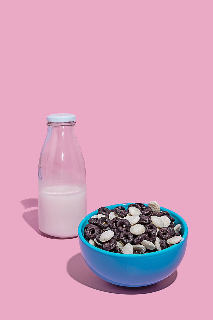 A vibrant photo featuring a blue bowl filled with chocolate cereal next to a bottle of milk against a pink background.