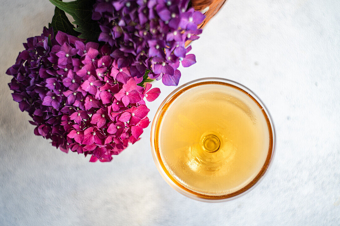 Blick von oben auf ein Glas mit einem frischen Getränk, das auf dem Tisch neben einem Strauß lila Hortensien in einem Korb mit brauner Tasche steht