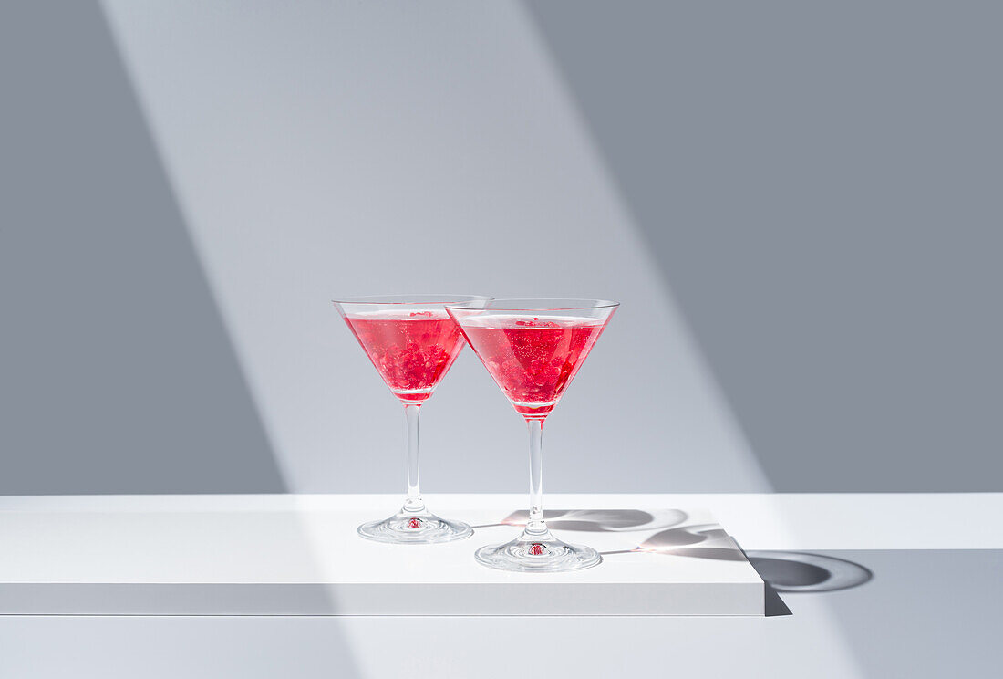 Mit roten Granatapfelcocktails gefüllte Gläser, die auf einer spiegelnden Oberfläche unter einem weichen Lichtstrahl platziert sind, wodurch symmetrische Schatten entstehen