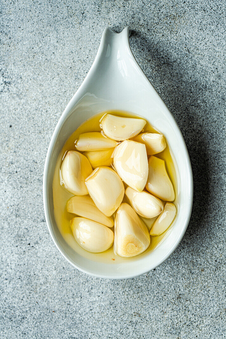 Top view of peeled and baked garlic cloves submerged in oil, showcased in a ceramic spoon against a light gray background
