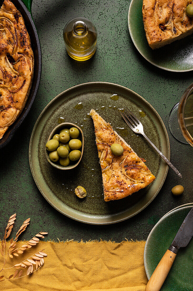 Draufsicht auf ein Stück Brot mit grünen Oliven, das mit einer kompletten im Ofen gebackenen italienischen Focaccia in einer Pfanne auf einem Esstisch mit Messer und Gabel zubereitet wurde