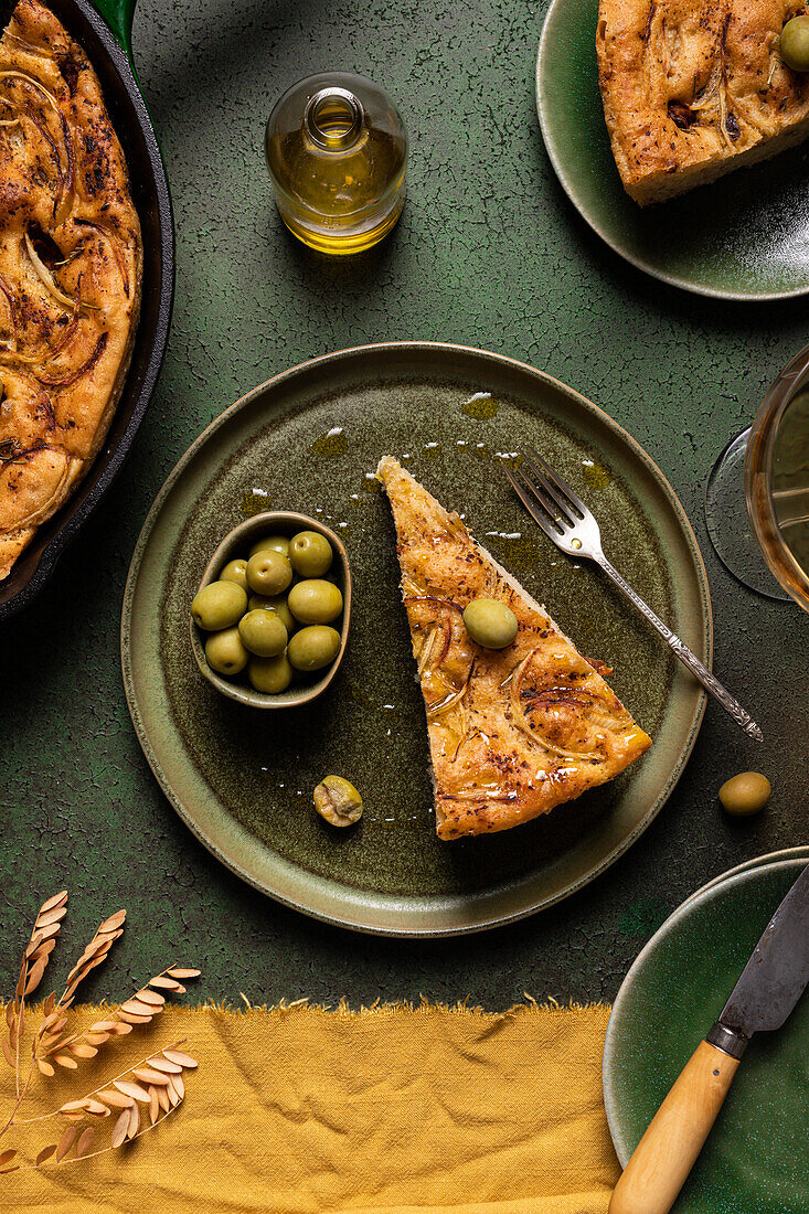 Top view of bread piece with green olives placed with complete oven baked Italian focaccia preparation in pan on dining table with knife and fork