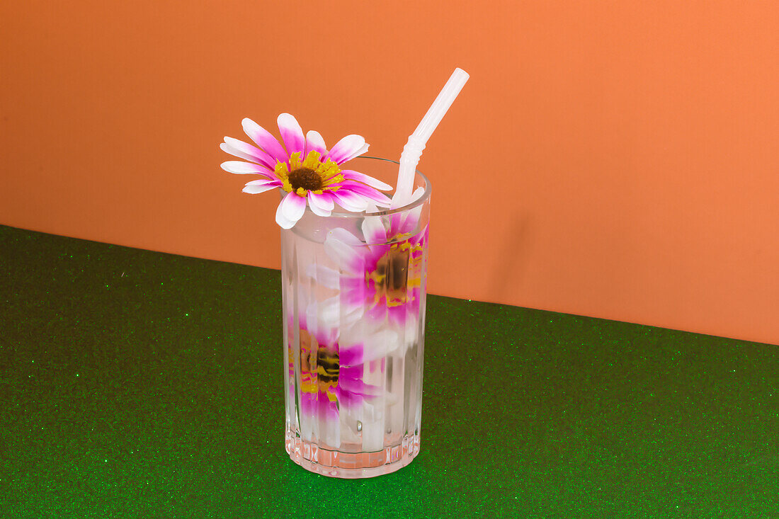 Transparent glass of refreshing cold drink decorated with pink flowers and straw placed on green surface against bright orange wall