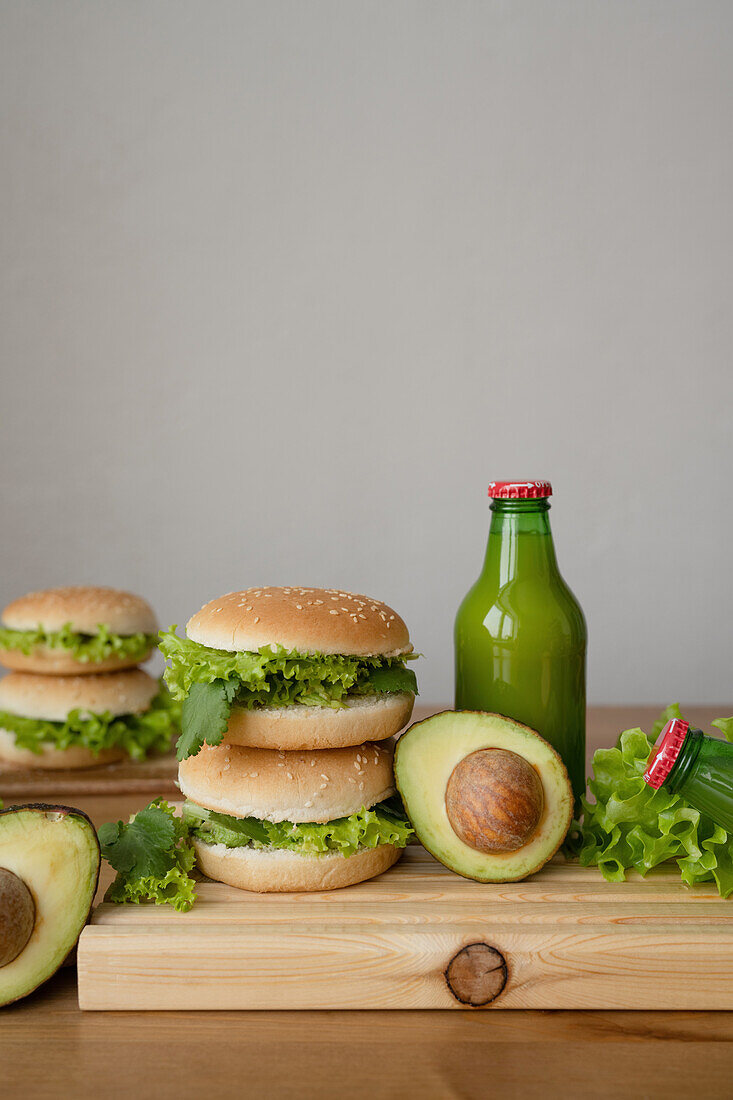 Tasty vegetarian burgers with fresh lettuce near avocado and bottle of juice on wooden cutting board against gray background