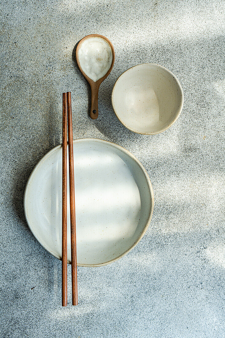 Draufsicht auf ein Keramikgeschirrset, bestehend aus Schüssel, Teller, Holzlöffel und Essstäbchen, auf einer grauen Fläche