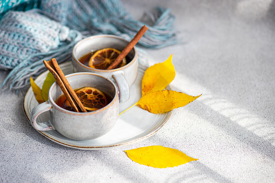 Cups of spiced tea adorned with cinnamon sticks, anise, and dried orange slices, nestled next to vibrant yellow leaves and a folded blue scarf on a soft gray backdrop