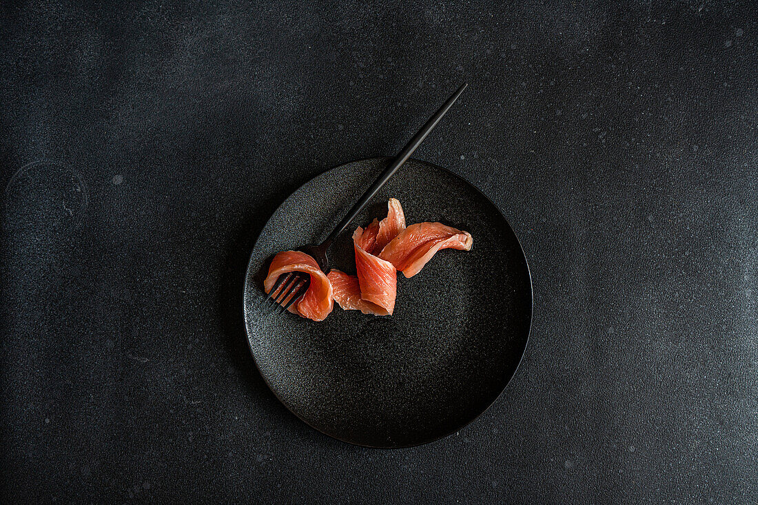 Draufsicht auf eine gesunde Lachsscheibe, serviert auf einem schwarzen Teller neben einer Gabel vor dunklem Hintergrund