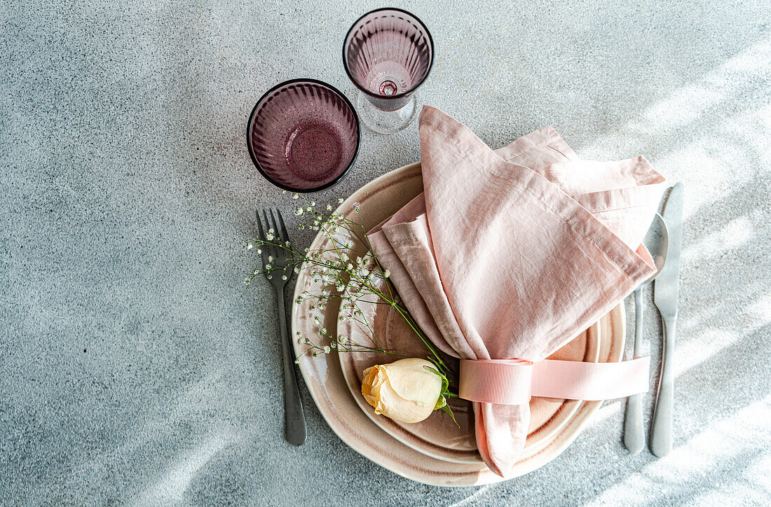A tastefully arranged Easter table setting featuring pastel-colored plates, napkins, and a delicate flower, accompanied by silverware and patterned glassware, all laid on a textured surface.