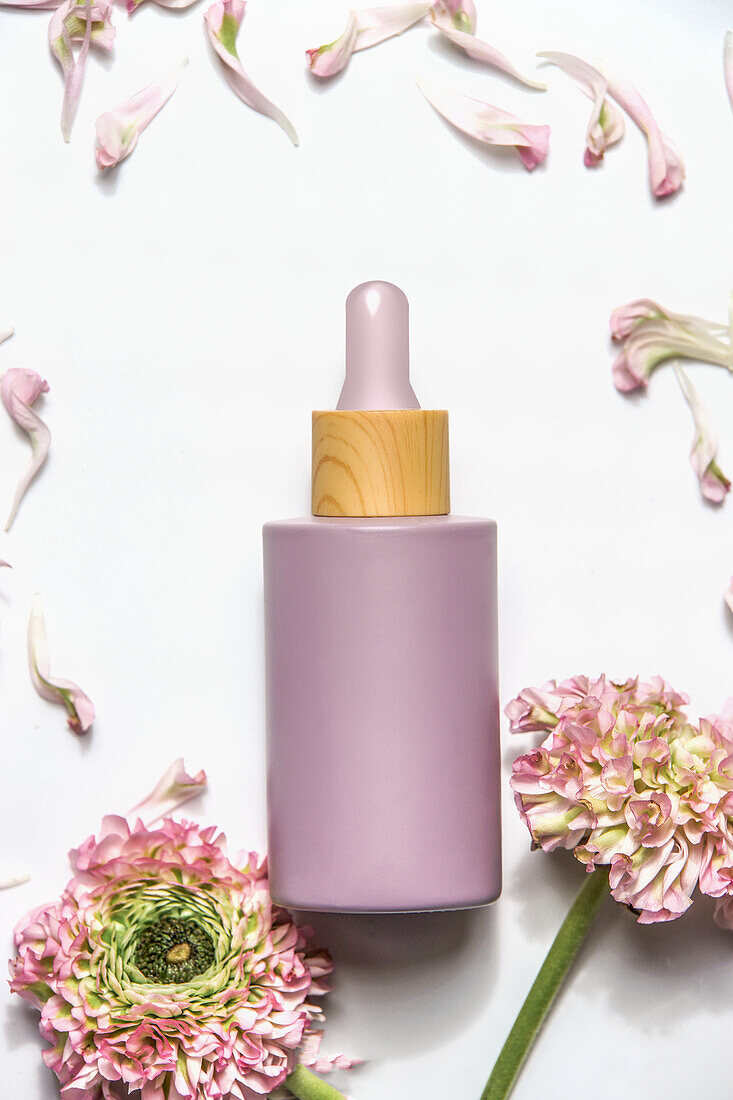 Pastel purple cosmetic bottle with pipette on white background with pink flowers , top view. Modern beauty product for skin care with copy space.
