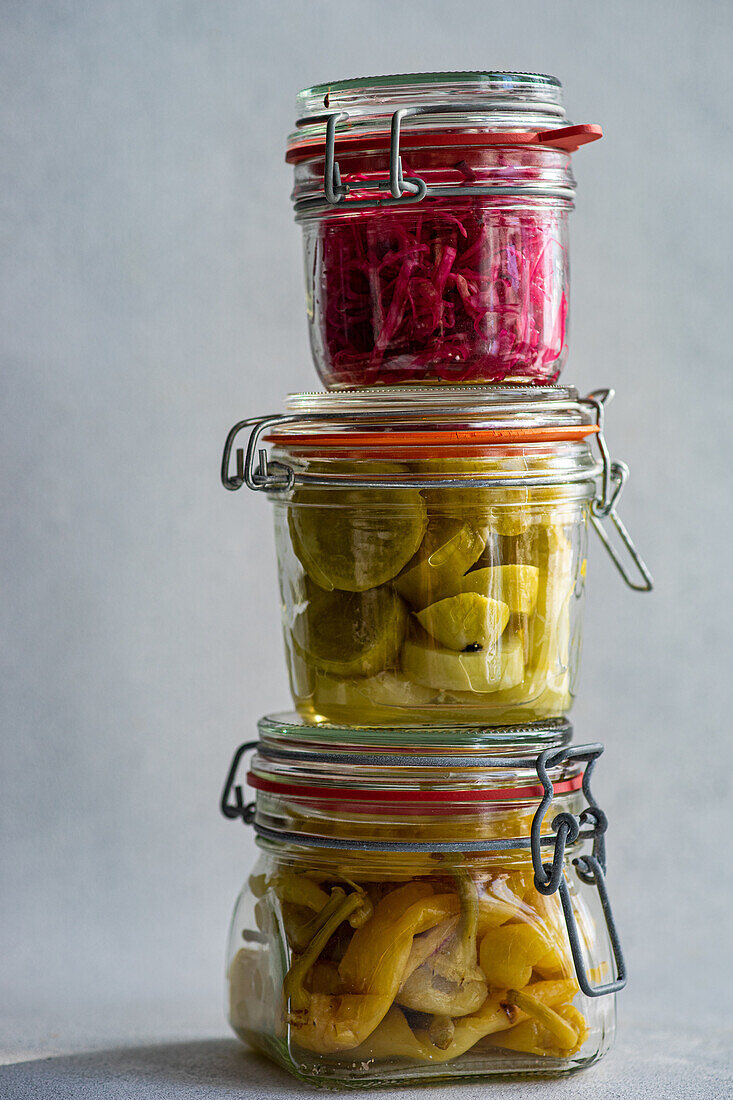 Three glass jars stacked atop each other, filled with fermented vegetables: red cabbage with beetroot, spicy peppers, and white cucumbers, showcasing a variety of vibrant colors and textures.