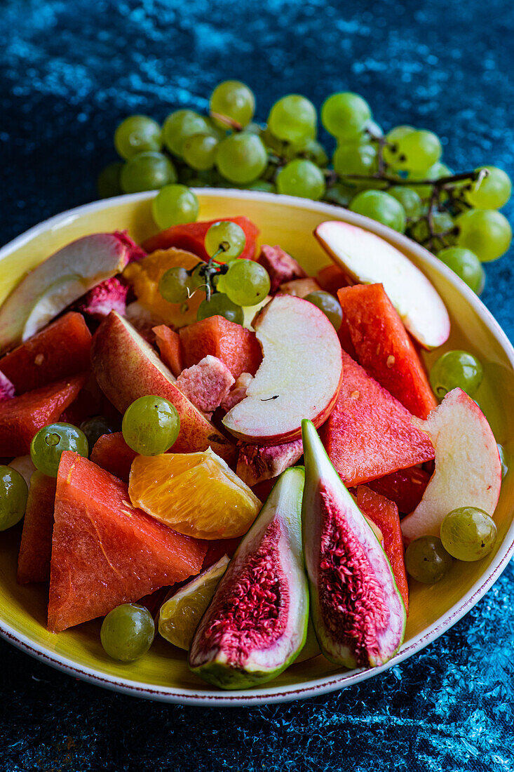 High angle of crop ceramic bowl of slices of different fruits placed on blue surface near blurred grapes