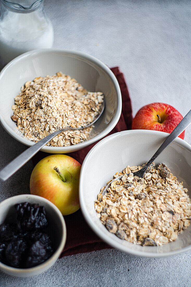 High angle of crop bowls with uncooked oatmeal placed on gray table near apples, milk jar and raisins