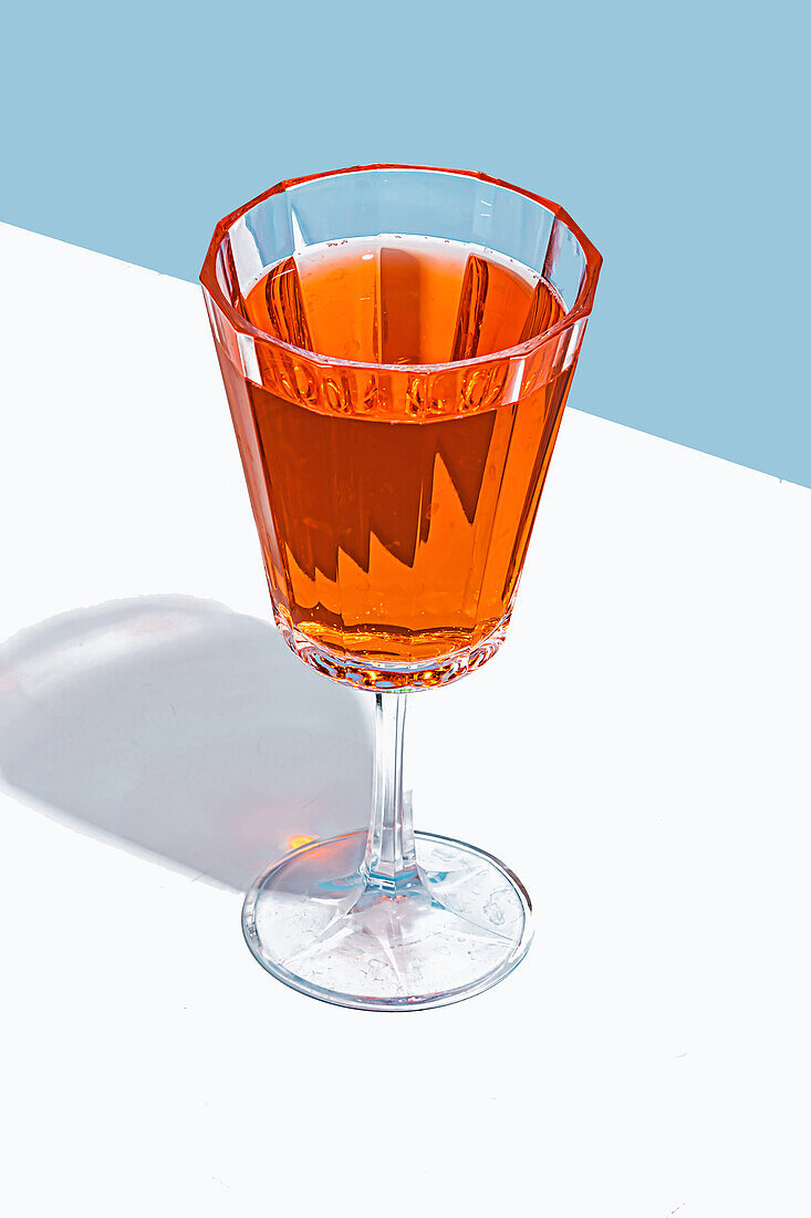 A clear, stemmed glass filled with an amber-colored liquid against a white surface with a blue background, casting a sharp shadow.