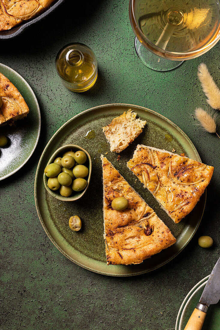 Draufsicht auf eine Scheibe gebackenes italienisches Focaccia-Brot mit grünen Oliven in einem Teller auf einem Esstisch mit Getränk auf grünem Hintergrund