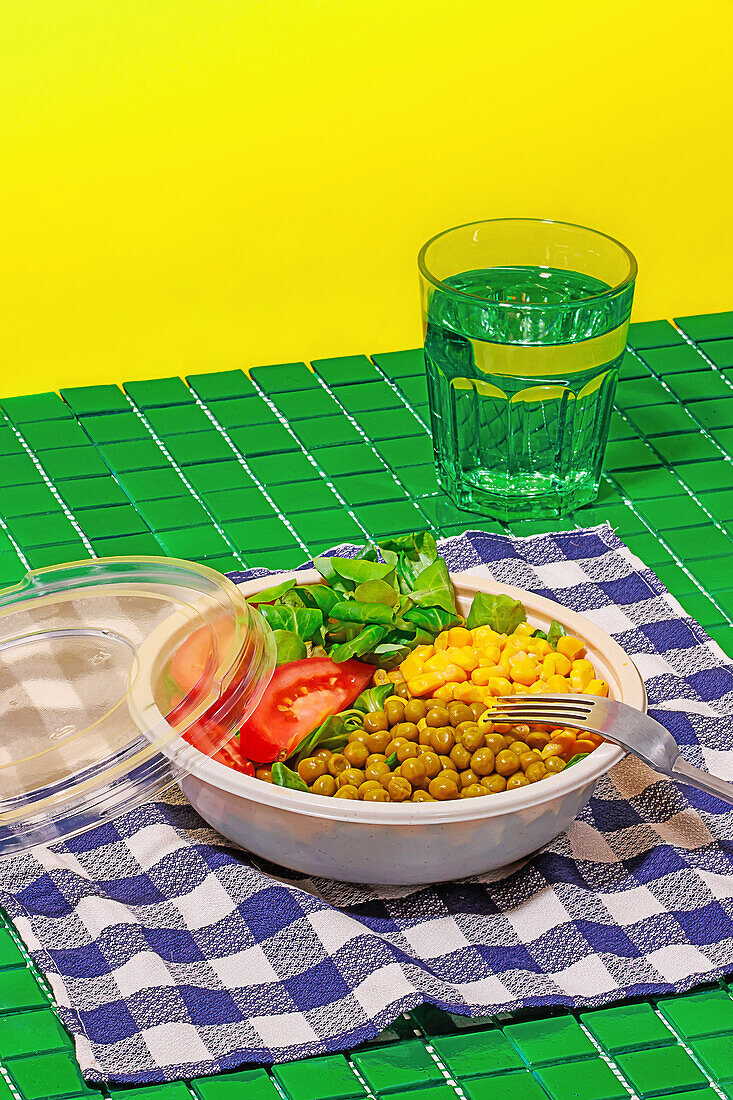 High angle of salad bowl with slices of tomato, spinach leaves, corn kernels and peas placed on napkin on green surface with fork near glass of water against yellow wall