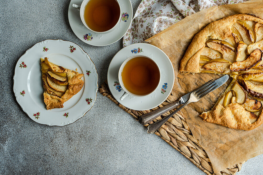 Top view of plates with pieces of apple pie and cups of tea near knife, fork and napkin on rustic tray against gray table