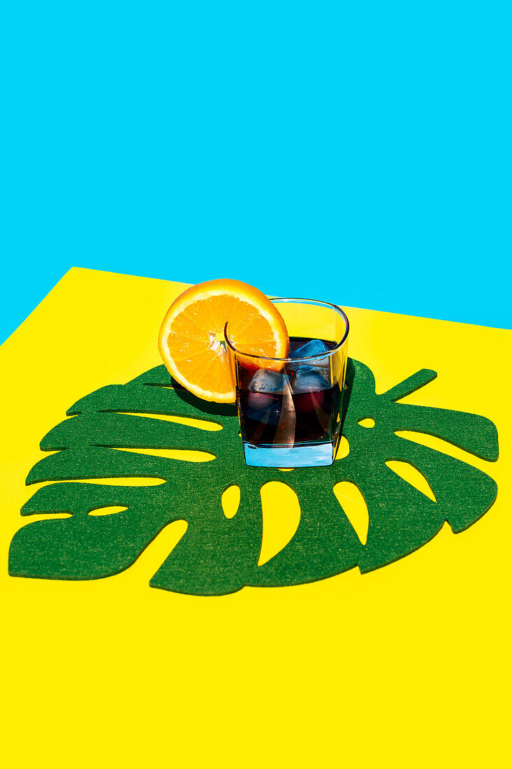 Kaltes Getränk mit Orangenscheibe im Glas auf Blattdekor über gelbem Hintergrund vor blauem Hintergrund im Studio