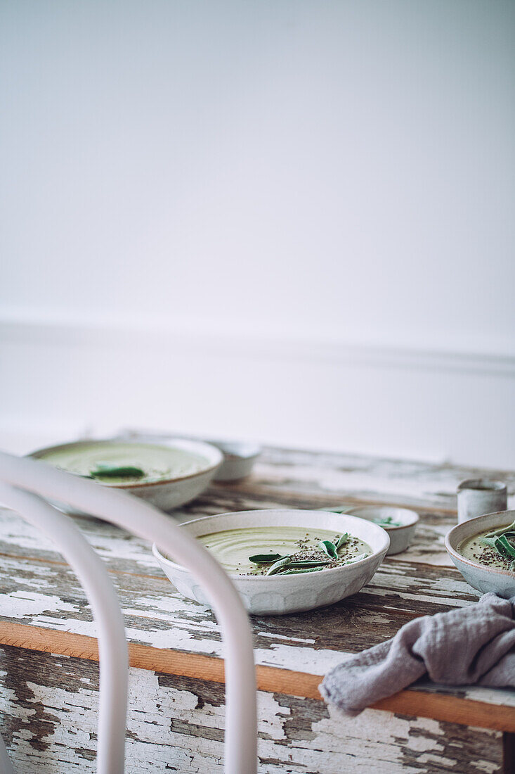 Ceramic bowls of green cream soup with sage served with seeds and herbs on wooden table with shabby surface against white background