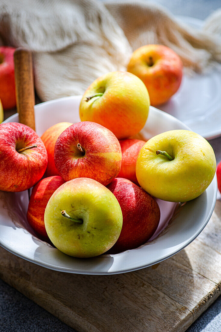 Draufsicht auf eine Sammlung von reifen, bunten Äpfeln, die auf einem rustikalen Holzbrett präsentiert werden, mit einem weißen Teller, einer Gabel und drapiertem Stoff, der eine heitere Küchenumgebung schafft