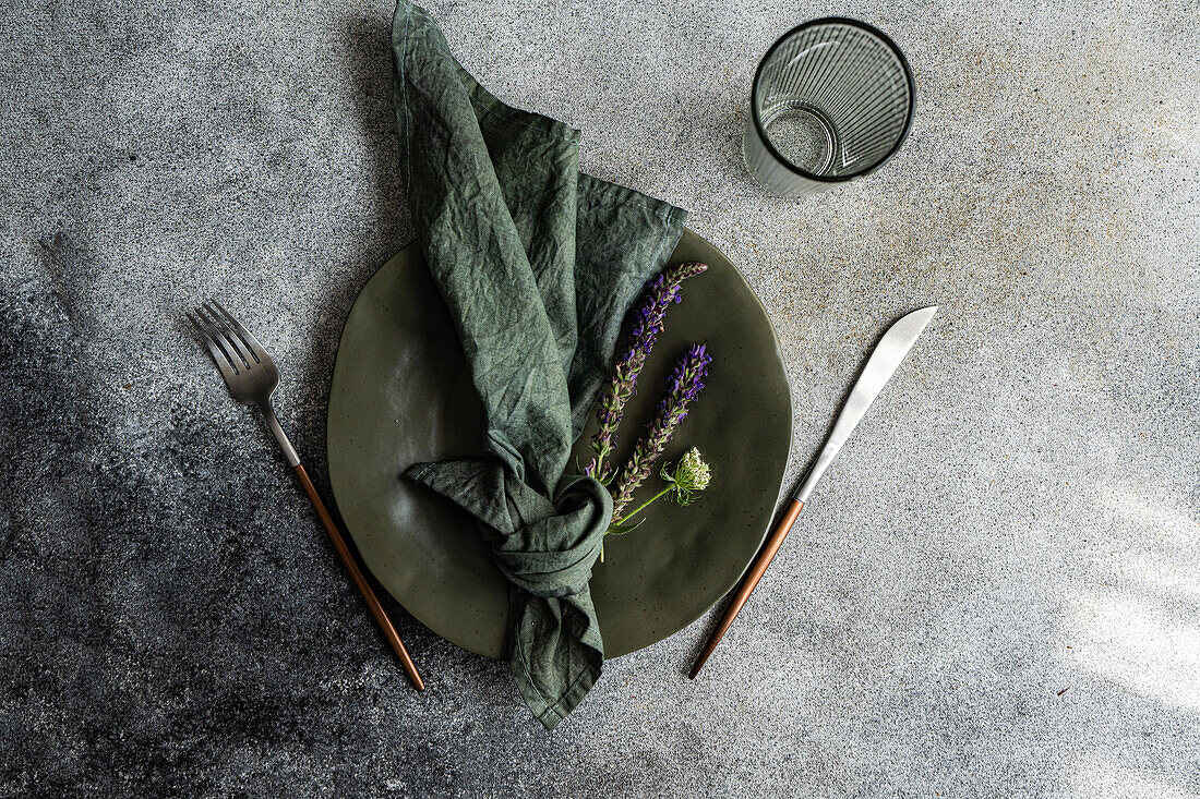 Draufsicht auf einen herbstlich gedeckten Tisch mit marmoriertem Teller, Serviette, Gabel und Messer und Lavendelblüten neben einem leeren Glas vor grauem Hintergrund