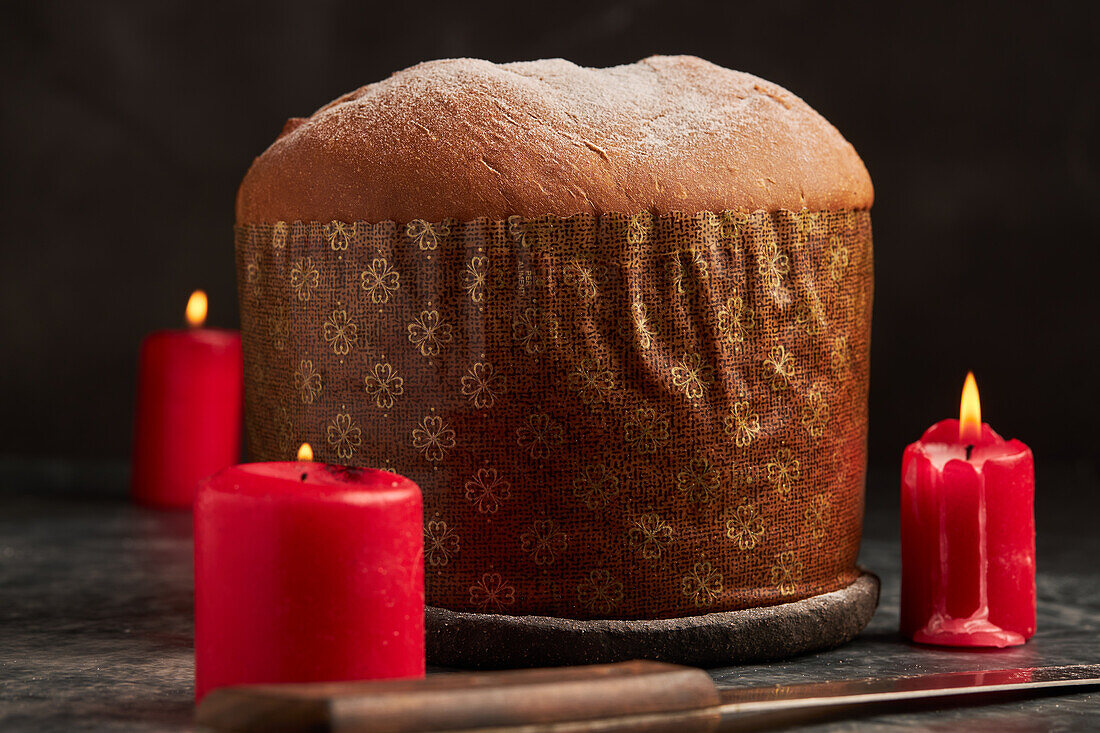 A traditional panettone bread, dusted with sugar, set against a dark backdrop with glowing red candles adding warmth to the scene