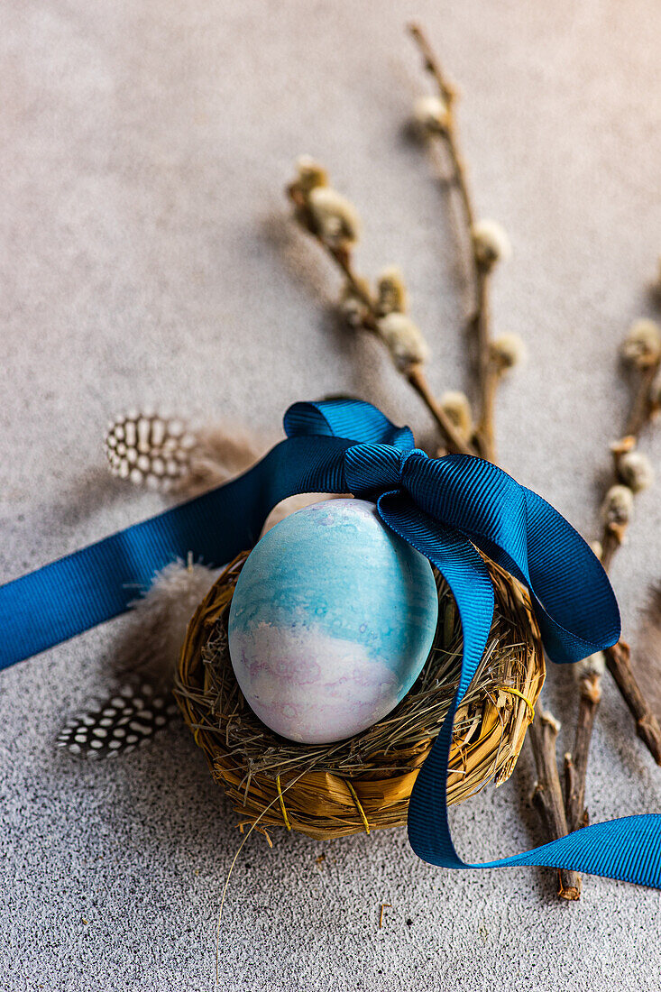 Close up of elegant Easter flatlay featuring a blue egg in a nest tied with a ribbon, accompanied by pussy willow branches and feathers on a textured surface