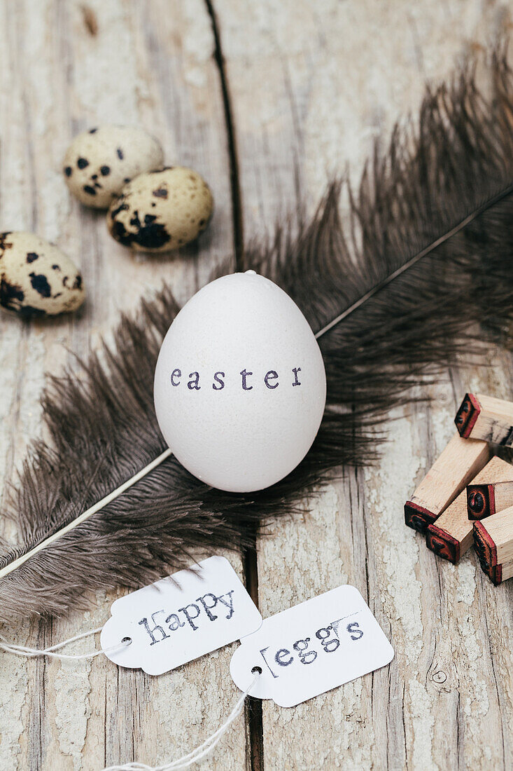 A white egg with "easter" stamped on it, surrounded by quail eggs, feathers, and "Happy eggs" tags on wooden surface.