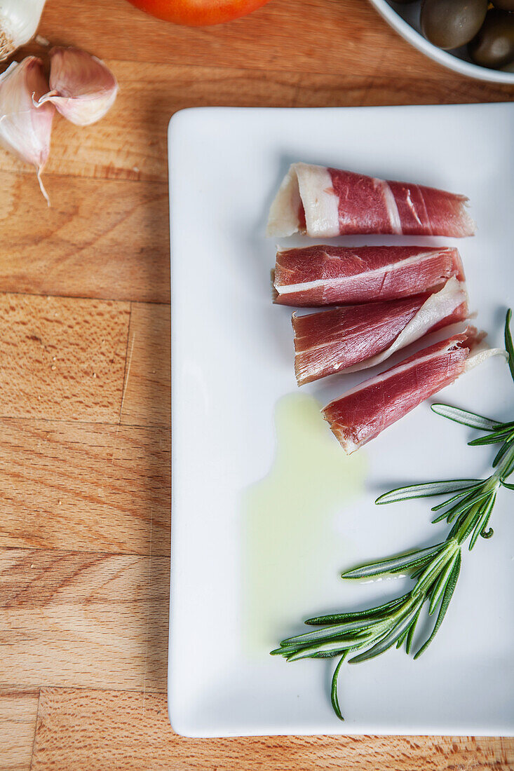 Sliced cured ham drizzled with olive oil beside fresh rosemary on a white plate, with garlic and a tomato in the background.