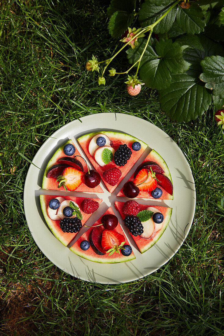 Top view of ingredients consisting of berries apple grapes plum peach placed on round cut slices in gray ceramic plate in sunlight on green grassy surface while preparing watermelon pizza