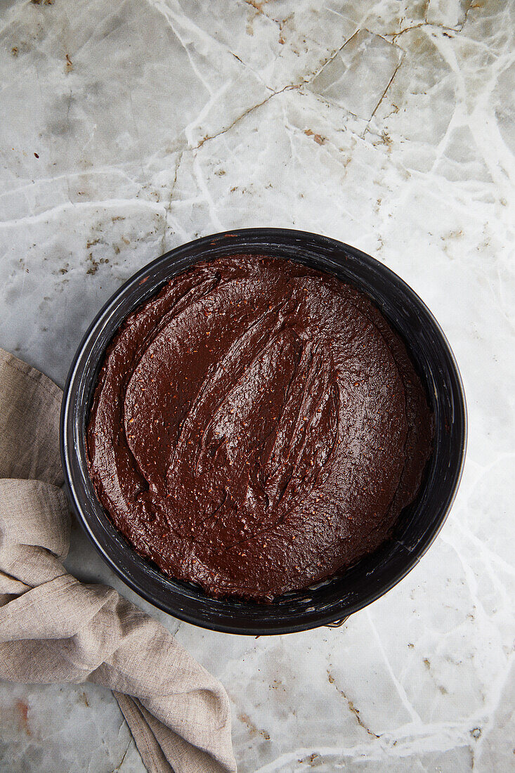 Blick von oben auf eine mit Schokoladenteig gefüllte Backform für Kuchen neben einem Tuch auf grauem Marmor