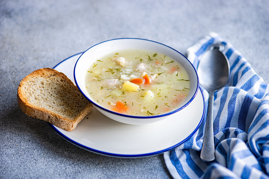 Freshly prepared chicken and vegetable soup in a white bowl with blue trim a piece of bread and a spoon on a blue striped cloth over a grey background