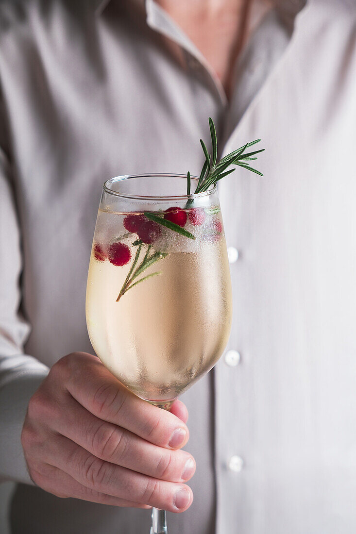 A hand holds a stemmed glass with a refreshing rosemary winter cocktail, garnished with cranberries and a sprig of rosemary.