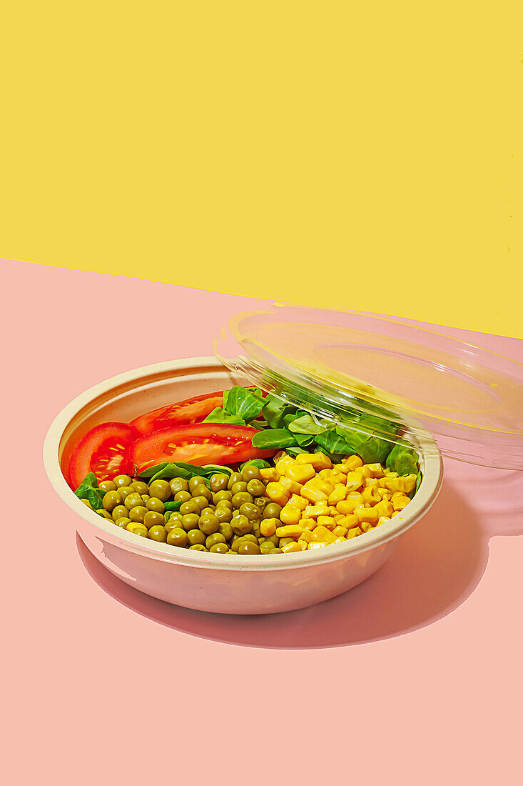High angle of salad bowl with slices of tomato, spinach leaves, corn kernels and peas placed on pink surface against yellow wall