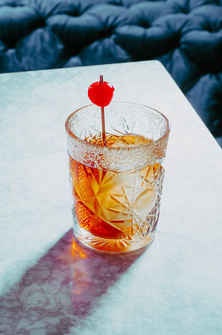 Blick von oben auf ein mit alkoholischem Old Fashioned Whiskey gefülltes Glas mit Eiswürfeln, garniert mit Kirschen, auf dem Tisch