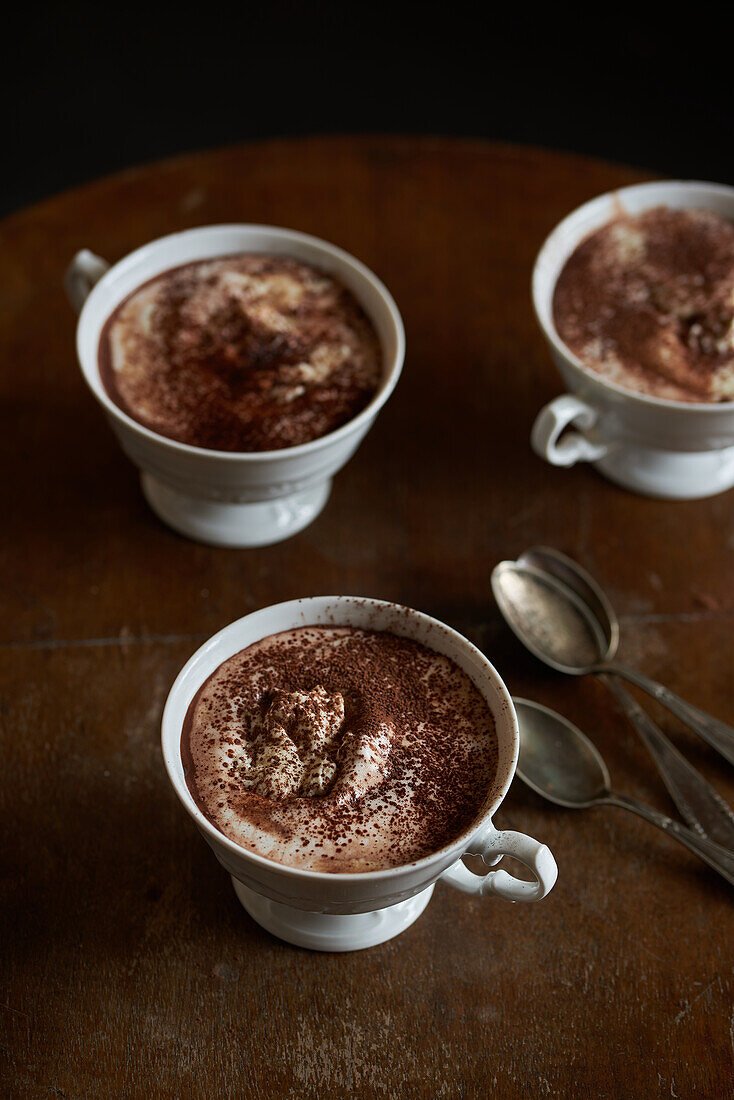 Two cups of hot chocolate topped with whipped cream and dusted with cocoa on a wooden table