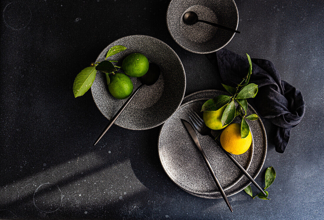 Artistic arrangement of ceramic dishes and fresh green lemons on a dark background.